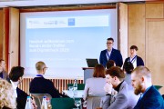Zwei Personen stehen in einem Raum und präsentieren etwas, auf einer Leinwand hinter ihnen steht: „Herzlich willkommen zum Bund-Länder-Treffen zum Digitalcheck 2025!“. Im Vordergrund sitzen weitere Personen und hören zu.