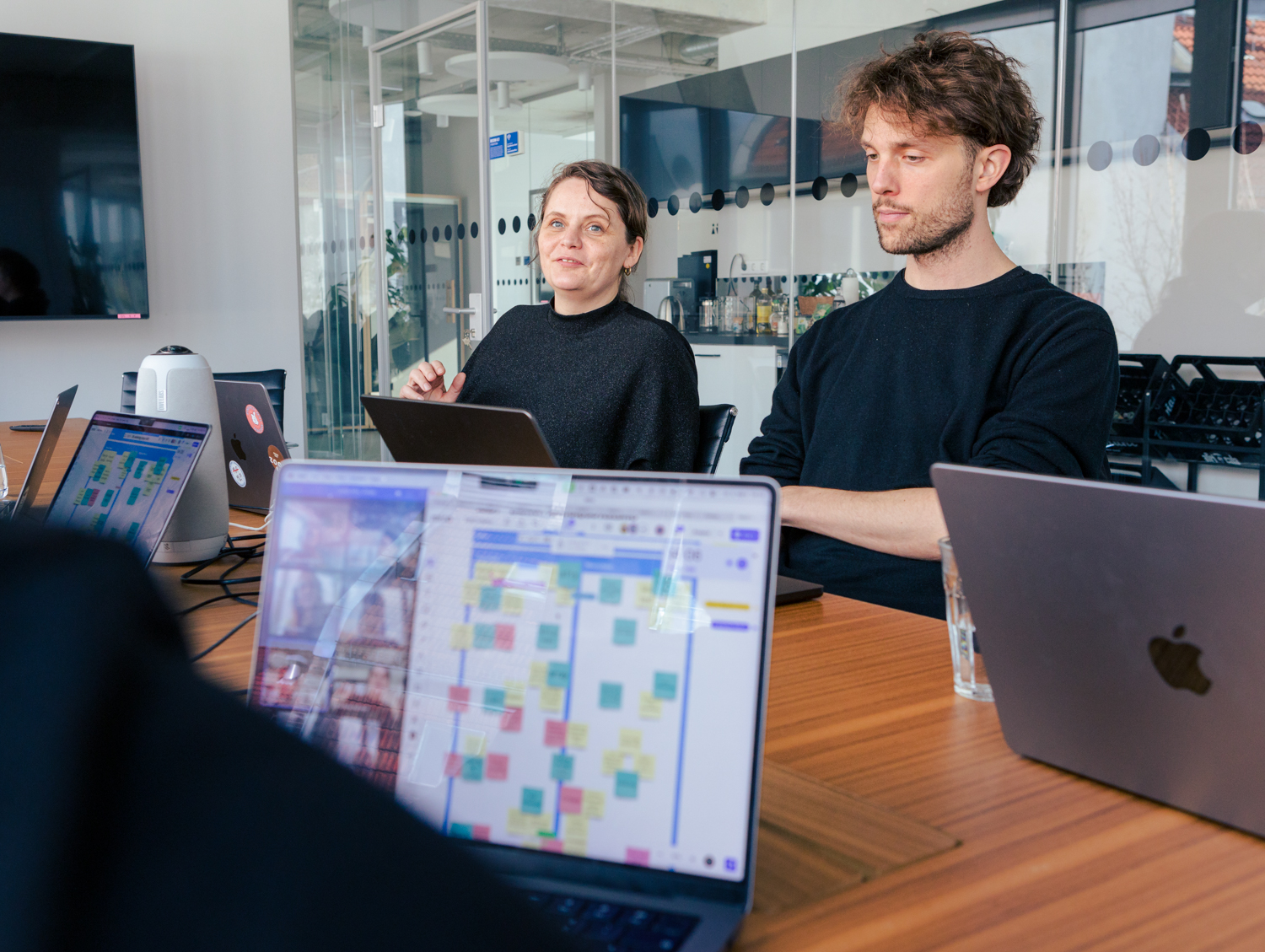 Mitarbeitende des DigitalService sitzen an einem langen Tisch mit aufgeklappten Laptops und diskutieren; auf den Laptops sind virtuelle Teilnehmende zu sehen und ein Board mit Notizzetteln